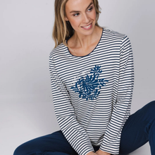 Load image into Gallery viewer, A woman wearing a navy and white horizontally striped long-sleeve top with a blue floral graphic on the front, paired with navy pants, seated against a light gray studio background.
