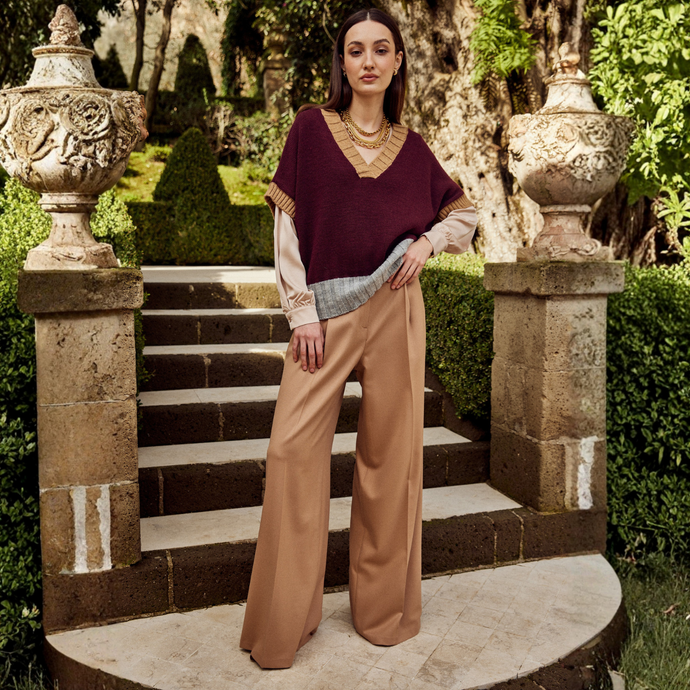 Woman in a burgundy top and beige pants standing on stone steps with decorative urns and greenery.