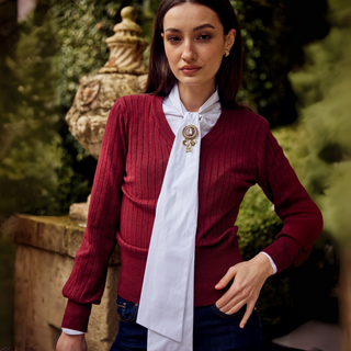 Woman wearing a red sweater over a white blouse with a decorative brooch, standing outdoors.