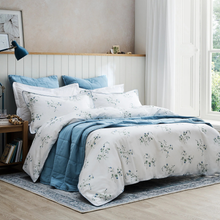 Load image into Gallery viewer, A neatly made bed in a bright bedroom featuring a white duvet cover and pillow shams with a delicate blue floral print, layered with solid medium-blue bedding and blue pillows. The room has light wood flooring, a window with white curtains, and a wooden headboard.
