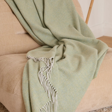 Load image into Gallery viewer, A sage green woven throw blanket with white fringe, draped over a beige armchair in a cozy indoor setting.
