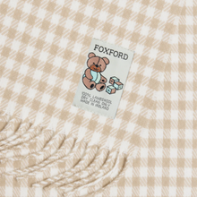 Load image into Gallery viewer, Close-up of a Foxford cream and beige checkered throw blanket with fringed edges and a sewn-on brand label featuring a teddy bear.
