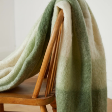 Load image into Gallery viewer, A soft, fuzzy throw blanket with a green and cream gradient draped over the back of a wooden chair in a bright indoor setting.
