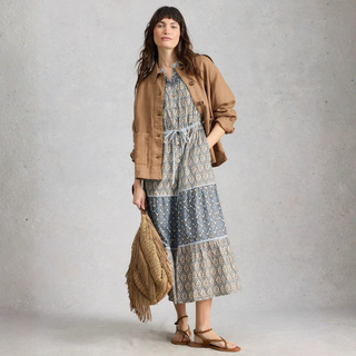 Woman wearing a patterned dress with a brown jacket and holding a fringed bag against a plain background