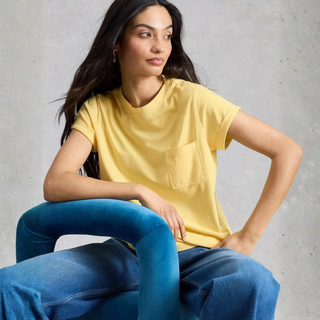 Woman wearing a yellow t-shirt and blue jeans sitting against a gray concrete wall.