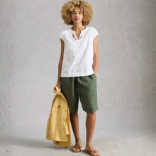 Load image into Gallery viewer, Woman wearing a white top and green shorts holding a yellow jacket against a gray background
