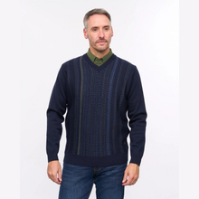 Load image into Gallery viewer, A man wearing a navy blue v-neck sweater with vertical textured knit stripes, layered over an olive green collared shirt and paired with blue jeans, photographed against a plain white studio background.
