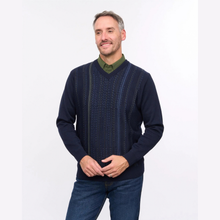 Load image into Gallery viewer, A man wearing a navy blue V-neck sweater with vertical textured knit panels over a green collared shirt and blue jeans, standing against a plain white studio background.
