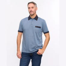 Load image into Gallery viewer, A man wearing a heathered blue short-sleeve polo shirt with a contrasting dark navy collar, sleeve cuffs, and a chest pocket, paired with dark blue jeans, against a plain white studio background.
