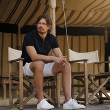 Load image into Gallery viewer, Man sitting on deck chair in navy polo and white shorts with trainers 
