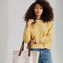 Load image into Gallery viewer, Woman wearing a yellow sweater and blue jeans, holding a beige tote bag with brown straps against a light gray background.
