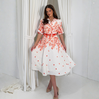 Woman wearing a white dress with red floral pattern standing in front of white curtains.