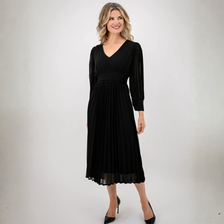A woman modeling a black midi dress with a V-neckline, long sheer sleeves, a defined waist, and a pleated skirt, paired with black heels against a light gray studio background.
