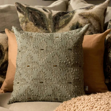 Load image into Gallery viewer, A square decorative throw pillow in a muted sage green with intricate floral embroidery and small metallic accents, styled on a sofa with other textured pillows in warm neutral and tan tones.
