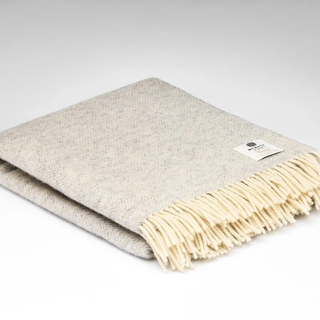 A folded woven throw blanket in a light beige and cream herringbone pattern with cream fringe, featuring a small white brand label on the corner.