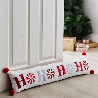 Decorative pillow with 'HO HO HO' text and red pom-poms on a wooden floor with a Christmas tree in the background.
