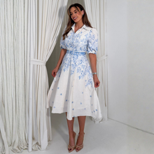 Load image into Gallery viewer, Woman wearing a white dress with blue floral patterns standing in front of white curtains.
