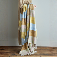 Load image into Gallery viewer, Person holding a striped blanket with yellow, blue, and beige colors on a wooden floor.
