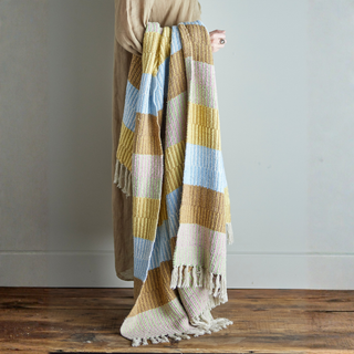 Person holding a striped blanket with yellow, blue, and beige colors on a wooden floor.