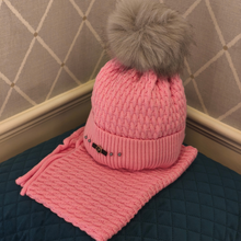 Load image into Gallery viewer, A pink knit beanie hat with a fluffy gray faux-fur pom-pom, shown with a matching pink knit scarf draped underneath on a quilted dark blue surface against a beige patterned wall.
