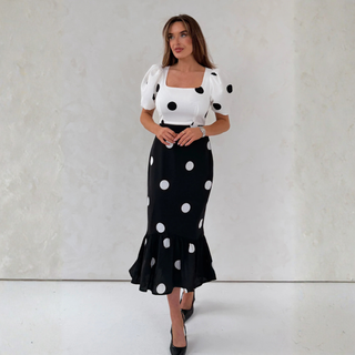 Woman wearing a white top with black polka dots and a black skirt with white polka dots against a plain background.