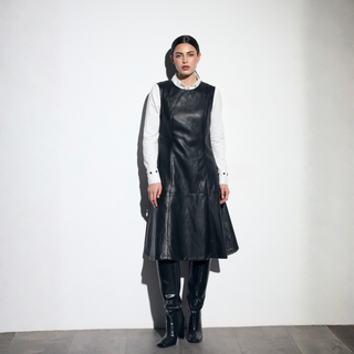 A model stands against a white studio wall wearing a black leather sleeveless dress over a white collared shirt, with black leather knee-high boots.
