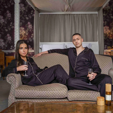 Load image into Gallery viewer, A man and a woman sitting on a sofa, holding a glass of prosecco, both while wearing the Jo Browne Luxury Unisex Bamboo Pyjamas. 
