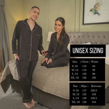 Load image into Gallery viewer, A man standing while a woman is sitting, both wearing  the Jo Browne Luxury Unisex Bamboo Pyjamas. A unisex size guide for the pyjamas is included in the image. 
