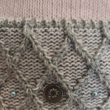 Load image into Gallery viewer, Close-up of a knitted garment or textile detail in light beige and gray, featuring a braided cable pattern, small pearl-like beads, and a round metallic button with a crystal center.

