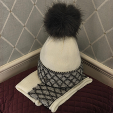 Load image into Gallery viewer, A white knit beanie hat with a large dark gray faux fur pom-pom, paired with a matching gray and white knit neck warmer, displayed on a quilted burgundy surface against a patterned wall.
