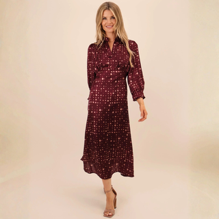 A woman modeling a long-sleeved midi dress in burgundy with a small gold polka dot pattern, set against a plain beige studio background.