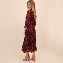 Load image into Gallery viewer, A woman modeling a long-sleeve midi dress in burgundy with a small pink and gold polka dot pattern, shown from a three-quarter back angle against a solid beige studio background.
