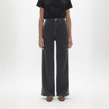 Load image into Gallery viewer, A model wearing a pair of dark gray wide-leg denim jeans, paired with a black graphic t-shirt and leopard-print flat sandals, standing against a plain white studio background.

