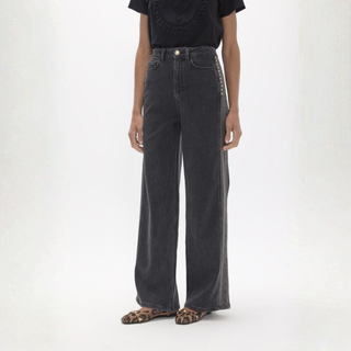 A model wearing a pair of dark gray wide-leg jeans with silver studs down the side, paired with a black t-shirt and leopard print flat shoes, against a plain white studio background.