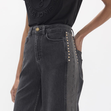 Load image into Gallery viewer, Close-up of a person wearing dark gray wide-leg jeans with gold-tone stud detailing along the side seams, paired with a black graphic T-shirt against a light gray studio background.
