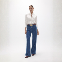 Load image into Gallery viewer, A model stands against a light gray studio background wearing a white long-sleeve button-up shirt with a small gold embroidered logo on the chest, blue wide-leg jeans, a brown belt, and brown pointed shoes.
