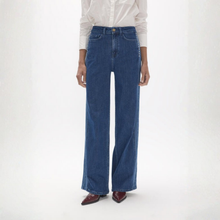 Load image into Gallery viewer, A model wearing a pair of high-waisted, wide-leg blue denim jeans with a white button-down shirt and dark brown pointed-toe mules, standing against a plain white studio background.
