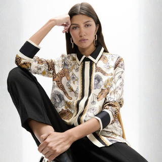 A model wearing a cream-colored silk-like button-up shirt with a detailed print of leopards, flowers, and stripes, paired with black trousers, set against a clean white background.