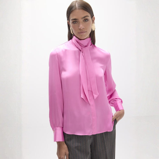 A woman modeling a bright pink long-sleeve silk blouse with a high neck and wide satin bow tie detail, paired with dark pinstripe trousers against a clean white studio background.