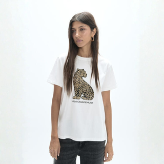 A woman modeling a white short-sleeve graphic T-shirt featuring a large leopard print on the front with the text 'OLA CASADEMUNT' underneath, paired with dark jeans against a plain white studio background.