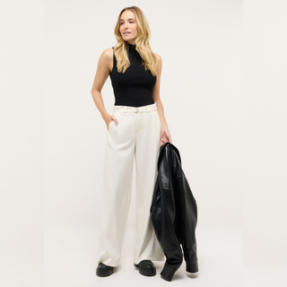 Woman wearing a black sleeveless top and white pants, holding a black leather jacket on a white background