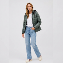 Load image into Gallery viewer, A woman modeling a dark green quilted puffer jacket over a white t-shirt, paired with light-wash blue jeans and white sneakers, against a plain light gray studio background.
