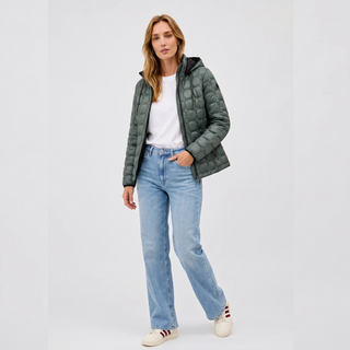 A woman modeling a dark green quilted puffer jacket over a white t-shirt, paired with light-wash blue jeans and white sneakers, against a plain light gray studio background.