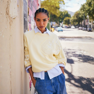 Woman wearing a light yellow sweater over a white shirt and blue jeans, leaning against a wall on a street.