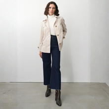 Load image into Gallery viewer, Woman wearing a beige trench coat, white turtleneck, dark blue jeans, and brown boots against a white background.
