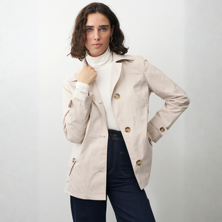 Woman wearing a beige trench coat over a white turtleneck and dark jeans on a plain background