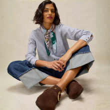 Load image into Gallery viewer, A model sits cross-legged on a light beige studio floor wearing a light blue and white striped button-up shirt with a green and black patterned scarf, blue denim shorts with turned-up hems, and brown suede clogs with gold-tone hardware.
