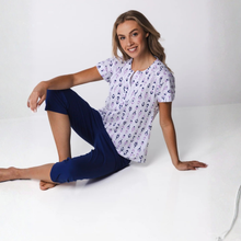 Load image into Gallery viewer, Woman sitting on a white surface wearing a patterned shirt and blue pants.
