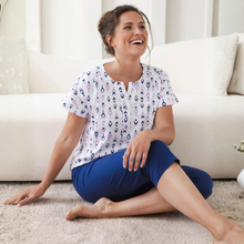 Load image into Gallery viewer, Woman sitting on the floor in a living room, wearing a patterned top and blue shorts.
