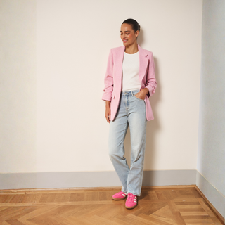 Person wearing a pink blazer, white shirt, light blue jeans, and pink shoes in a room with wooden flooring.